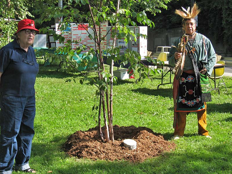 Jane Beecroft, in a red hat, stands beside a newly planted birch with Mississauga Elder Garry Sault in traditional regalia